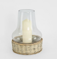 A clear glass hurricane lamp with a wide base, nestled into a woven natural wicker basket-like stand. The glass chimney is a classic, slightly flared shape, and the wicker base is a light, natural tone with candle inside