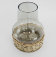 A clear glass hurricane lamp with a wide base, nestled into a woven natural wicker basket-like stand. The glass chimney is a classic, slightly flared shape, and the wicker base is a light, natural tone.