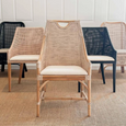 A dining chair with a frame made of light-coloured bamboo. The backrest is tall with a woven rattan insert and an arched top detail. The seat is a light, natural-coloured fabric cushion. The chair has armrests and a high back. Sitting with other dining chairs similar and in same collection.
