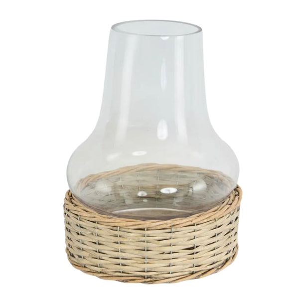 A clear glass hurricane lamp with a wide base, nestled into a woven natural wicker basket-like stand. The glass chimney is a classic, slightly flared shape, and the wicker base is a light, natural tone.