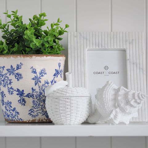 Hamptons-style home décor shelf with blue and white ceramic pot, white wicker container, decorative shell and photo frame.