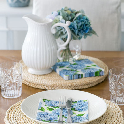 Hydrangea paper napkins styled on a coastal dining table with woven placemats, white stoneware and soft blue florals.