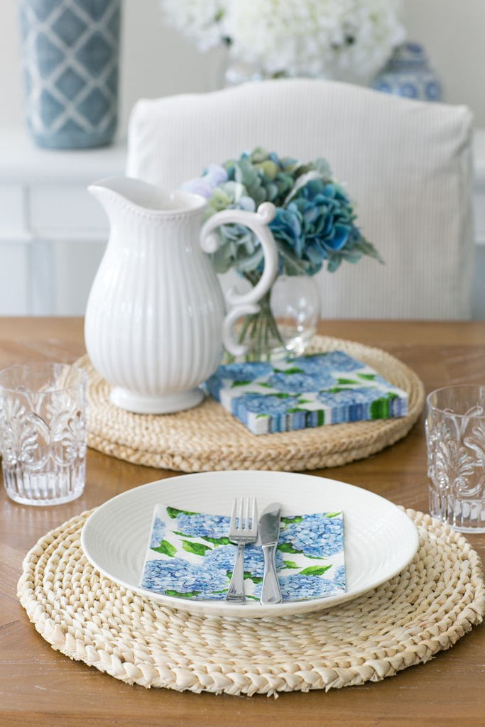 Hydrangea paper napkins styled on a coastal dining table with woven placemats, white stoneware and soft blue florals.