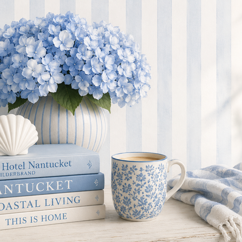 Blue hydrangeas in a striped ceramic vase beside coastal books, a shell ornament and blue floral mug on a light timber table.