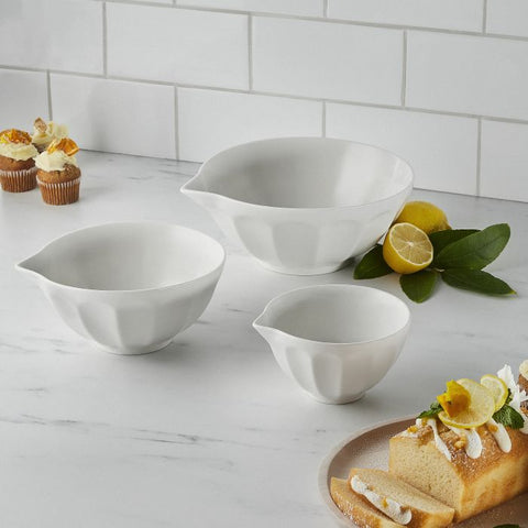 White ceramic mixing bowls styled on a marble kitchen bench with lemons and baked treats in a light coastal kitchen.