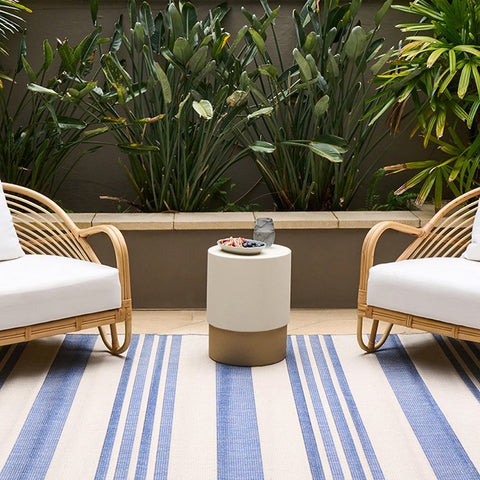 Blue and white striped indoor outdoor rug styled on a patio with wicker chairs in a coastal alfresco setting