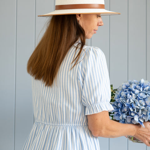 Woman wearing a white wide-brim hat and blue striped dress holding hydrangeas, styled in relaxed Hamptons coastal fashion