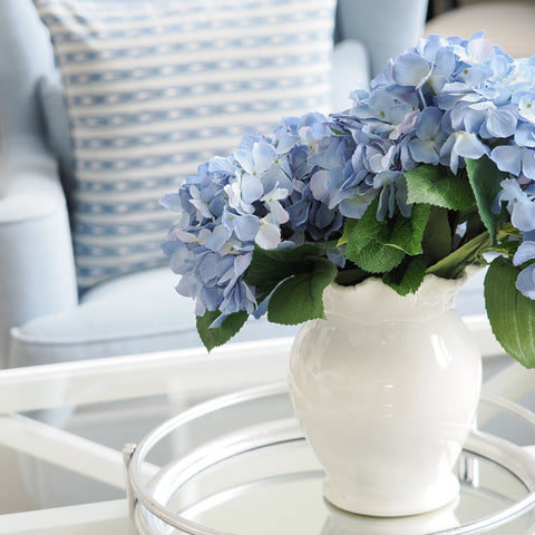 White ceramic vase with soft blue hydrangeas styled on a mirrored tray in a light Hamptons living room.