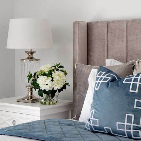 Bedroom styled with soft blue cushions, a white bedside table, glass lamp and layered textures in a calm Hamptons-style space.