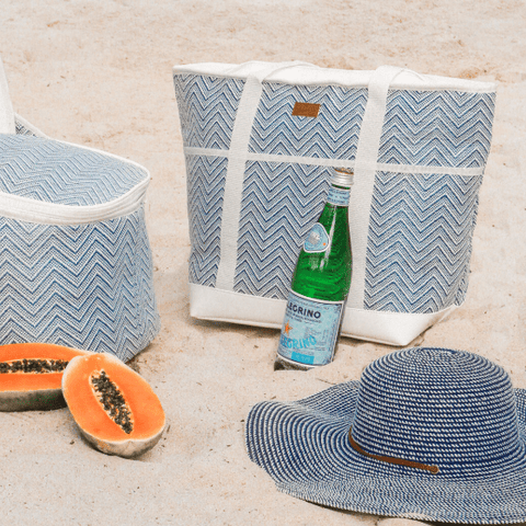 Coastal beach essentials including blue woven beach bag, picnic cooler and hat styled on sandy beach setting