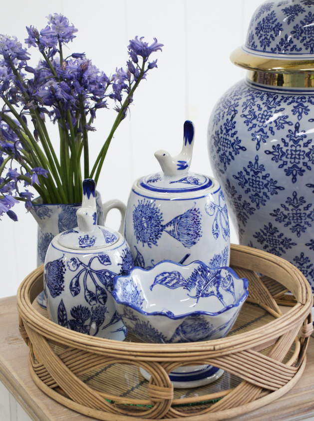 Blue and white ceramic ginger jars, lidded canisters, and a floral bowl styled on a rattan tray with fresh purple flowers in a coastal Hamptons setting.