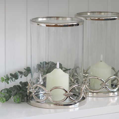 Glass hurricane candleholder with polished silver detailing and a soft pillar candle, styled on a white shelf.