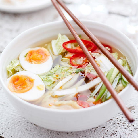 White ceramic serving bowl with noodle soup, eggs and vegetables, styled with wooden chopsticks on a light table.