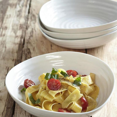 White ceramic serving bowls styled with pasta, cherry tomatoes and basil on a rustic wooden table.
