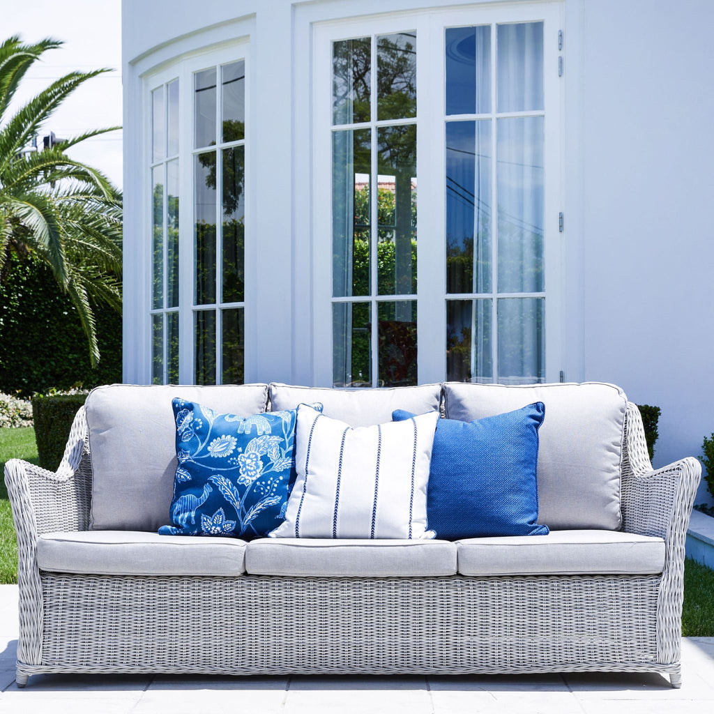 Hamptons-style outdoor wicker sofa with neutral cushions and blue accent pillows styled on a sunlit terrace with palm trees.