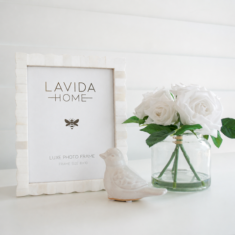 Sorrento Scallop Bone Photo Frame styled on a white shelf beside white faux roses in a glass vase and a ceramic bird ornament for relaxed coastal styling.