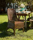 A natural brown rattan dining chair with a high, slightly curved backrest and armrests. The back and seat are both made from tightly woven rattan, and the chair has a classic, elegant silhouette.