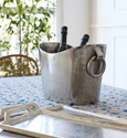 Antique-style silver wine bucket with large ring handles, featuring a curved rim and vintage finish, styled with champagne bottles on a dining table.