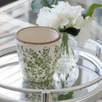 Decorative pot with green floral pattern on a glass table with a small vase of white flowers.