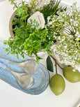 A coastal tabletop vignette with fresh greenery, soft blue serving spoons, and green citrus accents.