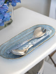 Coastal Hamptons-style blue ceramic serving platter with gold-edged detail, paired with white-handled salad servers on a bright white dining table.