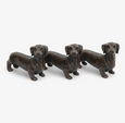 A set of three small, brown dachshund dog-shaped resin figurines. Each figurine has a different pose, and they are designed to be placed under a pot to elevate it.
