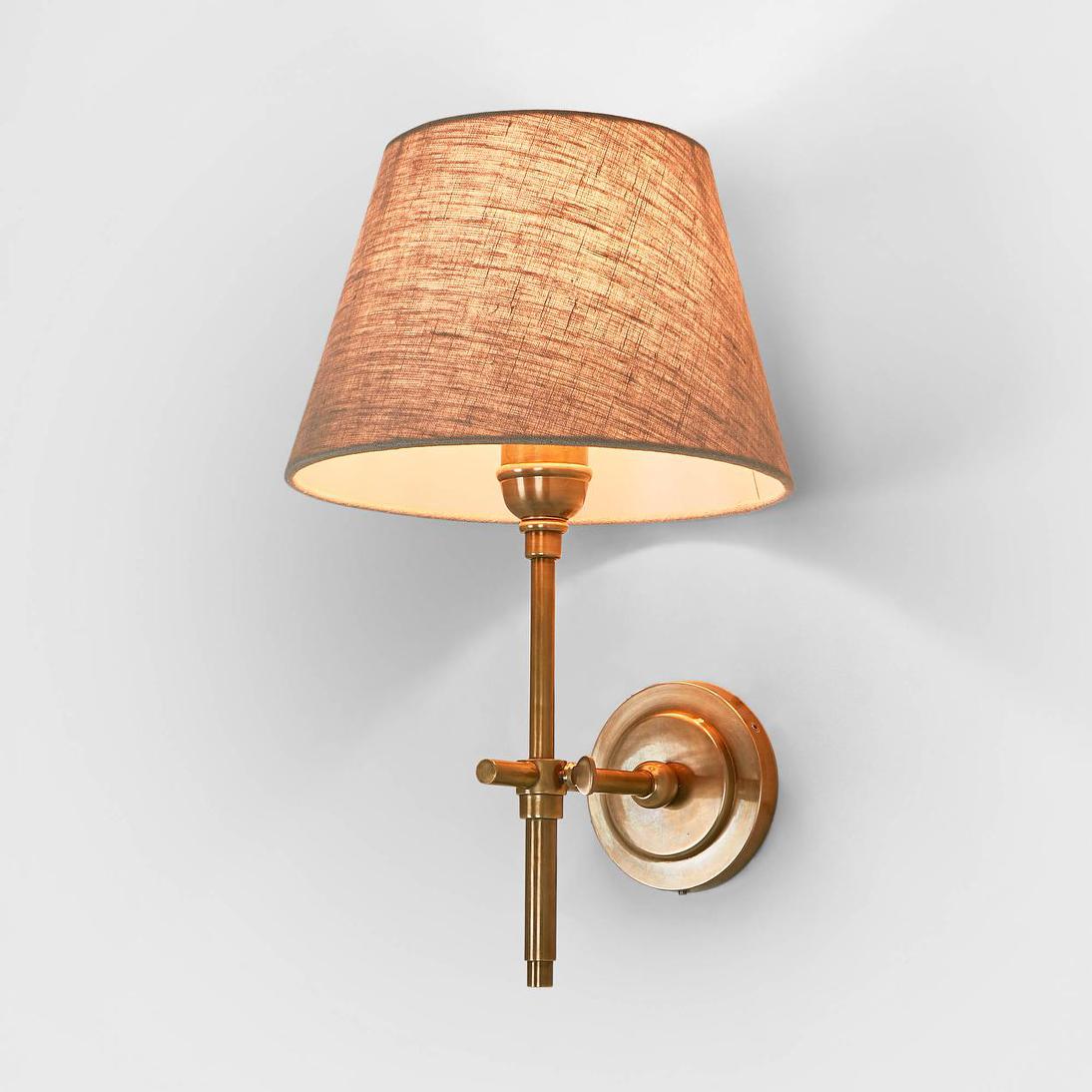 Dino Wall Light in antique brass with a round wall plate and slim stem, shown with a natural linen shade