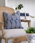 Eastport navy paisley cushion styled on a classic cane-back armchair with a soft upholstered seat, set against a bright white coastal bookshelf vignette.