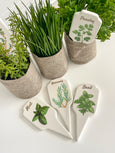Herb garden stakes set with illustrated ceramic markers for thyme, parsley, rosemary, mint and basil styled in linen pots on a white surface.