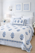 Hamptons-style bedroom featuring a white quilted coverlet with blue floral motifs, styled with layered pillows, soft blue walls and a natural jute rug.