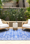 Blue and ivory flatweave outdoor rug featuring an elegant scroll pattern, styled in a Hamptons-inspired patio with rattan chairs and lush greenery.
