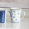 A tall ceramic mug with a white body. It features a scattered pattern of small blue roses and a detailed navy blue lace-like border along the top and bottom rims. The handle and interior are solid white.