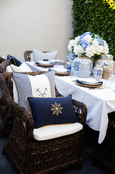Navy ship’s wheel cushion displayed on wicker seating beside a Hamptons-style outdoor dining table.