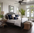 Hamptons-style coastal bedroom with navy and white cushions, dark timber bed frame, woven bench seat and palm accents, styled with French doors overlooking the water.
