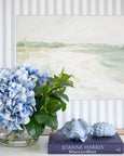 Blue hydrangea arrangement in glass vase styled with coastal shell décor and book, soft Hamptons beach artwork backdrop