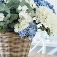 Blue and white faux hydrangea bouquet styled in a woven rattan basket on a timber dining table.