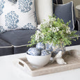 A styled living room scene featuring a dark gray sofa with decorative throw pillows, a white coffee table, and a woven tray holding a ceramic planter with greenery and white flowers.