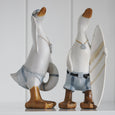 Two decorative duck figurines standing on a white surface against a light gray paneled background. One duck wears light blue denim shorts and a life preserver, while the other wears matching denim shorts with a black belt and holds a white surfboard with gold stripes.