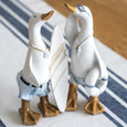 Two decorative white duck figurines standing on a striped white and navy table runner, one holding a white surfboard with tan stripes, against a blurred wooden floor background.