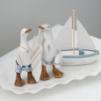 A decorative figurine set featuring two white geese dressed in blue shorts and gold necklaces, one holding a white surfboard and the other standing beside a small white sailboat with blue trim, all arranged on a white scalloped platter against a soft gray background.