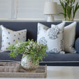 A styled living room scene featuring a navy blue sofa with decorative throw pillows, a white table lamp, and a white wall with shutters in the background. In the foreground, a wicker tray holds a ceramic vase filled with green foliage and small white flowers.