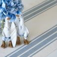 Two white decorative swan figurines wearing blue swim trunks and gold-toned sandals, one holding a white striped surfboard, styled on a blue-and-white striped runner with blue hydrangeas in the background.