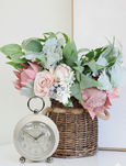 Ivory vintage alarm clock styled beside a wicker basket filled with soft pink roses, protea and eucalyptus on a white surface.