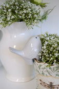 Small white ceramic pot sitter bird with sand-toned belly perched on the rim of a decorative pot filled with greenery.