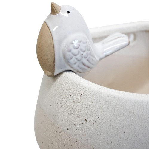 Small white ceramic pot sitter bird with sand-toned belly perched on the rim of a decorative pot filled with greenery.
