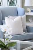 Light blue Hamptons style armchair styled with a white velvet cushion and striped cushion, paired with a white side table and orchid on a glass coffee table in a coastal living room.