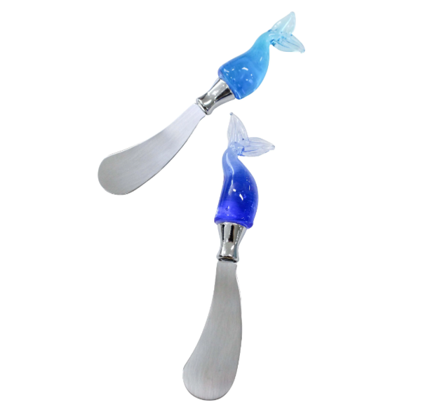 A set of two butter or cheese spreaders. Each spreader has a polished, silver stainless steel blade and a handle made of a translucent whale tail in shades of blue blown glass. One handle is a lighter blue and the other is a darker blue.