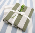 A tablecloth folded neatly, showing a wide vertical stripe pattern in alternating shades of light blue and white. The fabric has a relaxed, slightly textured finish. The tablecloth is tied with a white ribbon and has a brown product tag.