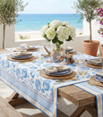 Coastal Hamptons-style outdoor dining table set with blue seashell tablecloth, woven placemats, hydrangea centrepiece and ocean view backdrop.