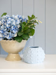 Cream ceramic pedestal urn styled with blue hydrangea stems beside a soft blue textured ceramic vase on a shelf, styled in a classic Hamptons coastal interior.